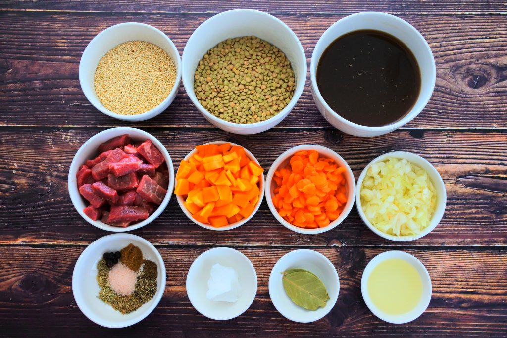 An overhead image of bowls of ingredients including lentils, quinoa, beef, beef bone broth, carrots, diced onion, diced butternut squash, light olive oil, bay leaves, coconut oil, Himalayan salt, black pepper, cumin, dried oregano, dried basil, dried rosemary, fennel seeds, rubbed sage, celery seeds, and juniper berries