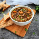 Close up image of a bowl of lentil and quinoa beef stew on a wood plank with pieces of bread on the side