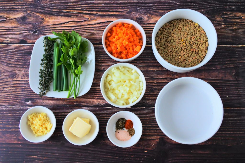 An overhead image of an assortment of bowls containing the ingredients for a coconut lentil stew including various seasonings, dry lentils, butter, minced garlic, chopped onion, chopped carrots, parsley, thyme, chives and coconut milk.