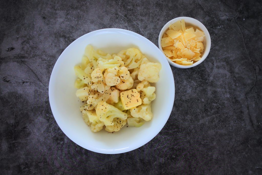 An overhead image of a bowl of steamed cauliflower florets with a pat of butter and a sprinkle of salt and black pepper on it and a smaller bowl of shaved Parmesan cheese to the top right of it.