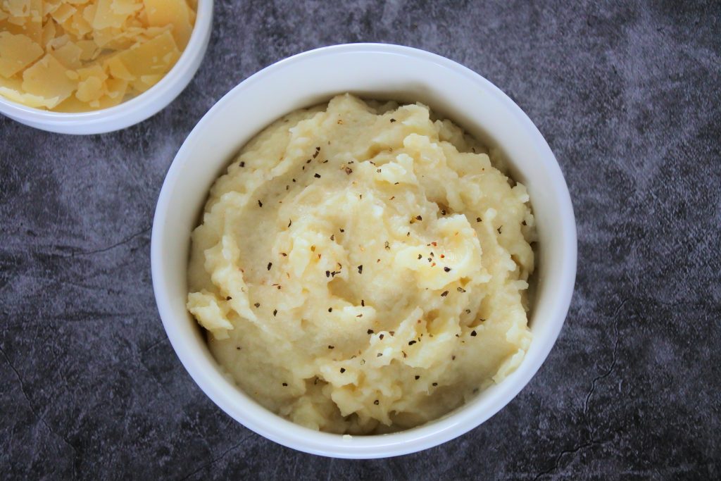 An overhead image of a bowl of mashed cauliflower with a scattering of black pepper on top and a bowl of shaved Parmesan cheese in the top left corner.
