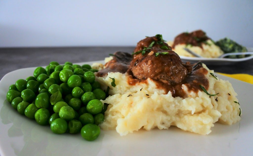 A close up image of a serving of mashed cauliflower topped with two Salisbury meatballs in gravy and a side of sweet peas with a second plated meal in the background.