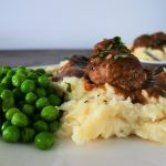 A close up image of a serving of mashed cauliflower topped with two Salisbury meatballs in gravy and a side of sweet peas with a second plated meal in the background.