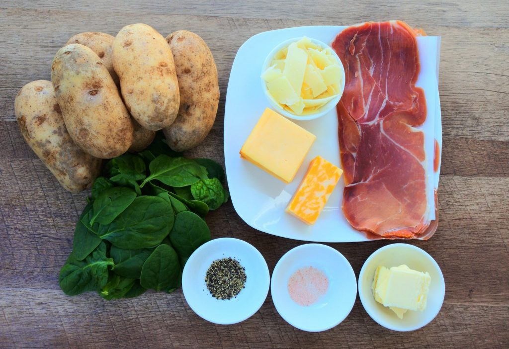 An overhead image of ingredients for next level mashed potatoes including potatoes, spinach, cheeses, salt, pepper, butter and prosciutto