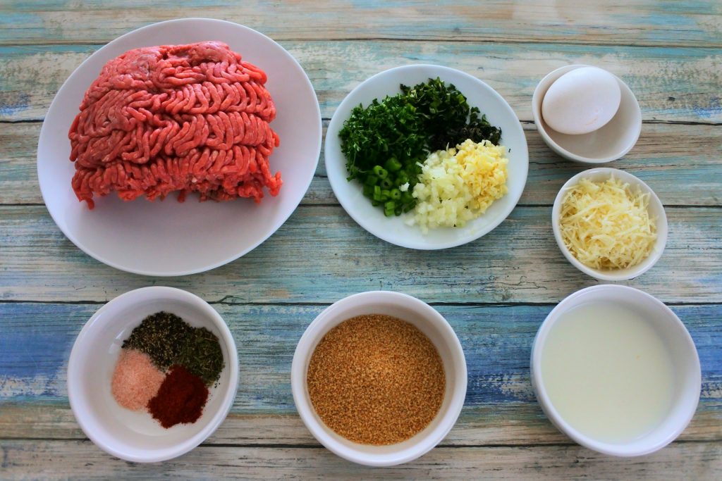 An overhead image of bowls of ingredients for meatballs including: ground beef, fresh herbs and spices, whole wheat breadcrumbs, milk, cheese and an egg.