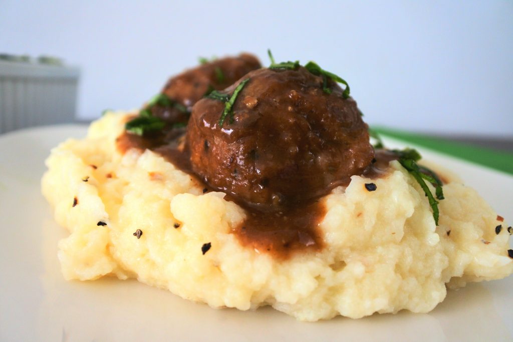 A close up image of a serving of mashed cauliflower topped with two Salisbury meatballs in gravy.