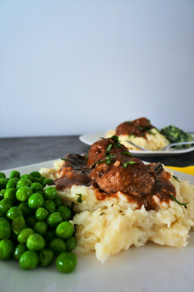 A vertical image of a serving of mashed cauliflower topped with two Salisbury meatballs in gravy and a side of sweet peas with a second plated meal in the background.