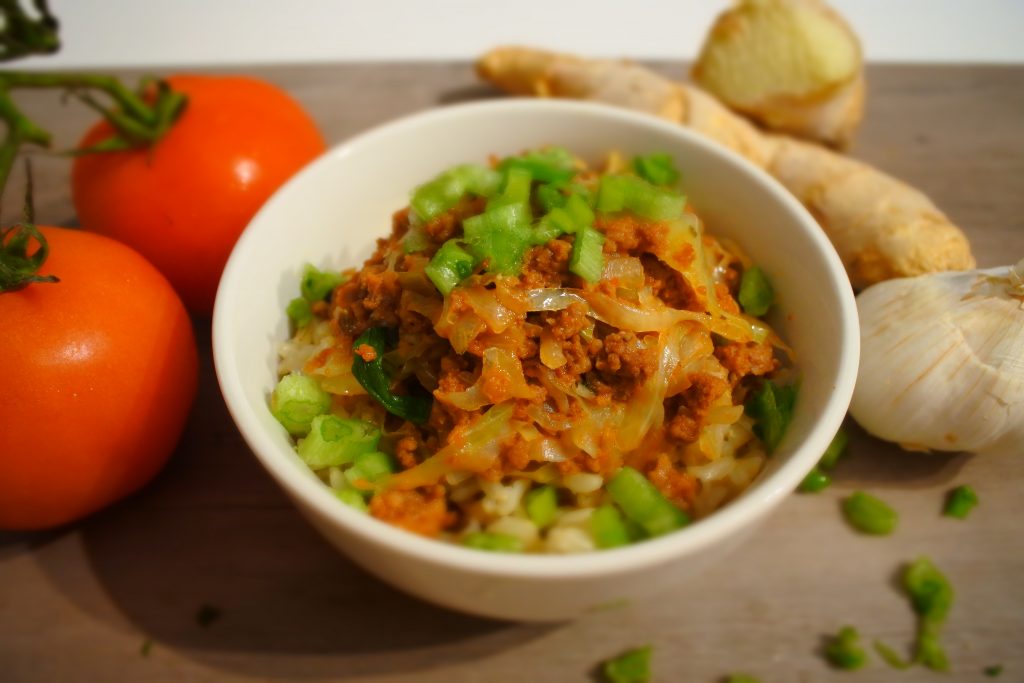 A tilted head on image of a bowl of brown rice topped with minced meat and cabbage and garnished with sliced chives, surrounded by fresh tomatoes, ginger, a head of garlic and a scattering of chopped chives.