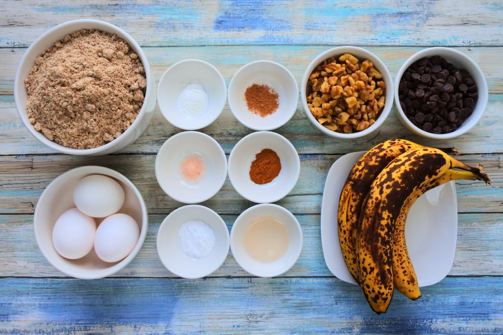 An overhead image of dishes containing ingredients for banana nut dark chocolate muffins including almond flour, eggs, bananas, baking powder, baking soda, nutmeg, cinnamon, salt, vanilla extract, chopped walnuts and dark chocolate chips.