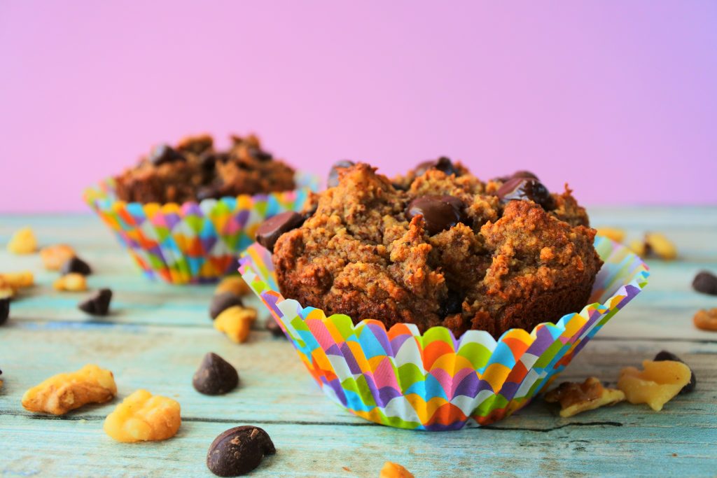 A close up image of a banana nut dark chocolate muffin in a colorful paper liner surrounded by dark chocolate chips and walnuts