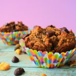 A close up image of a banana nut dark chocolate muffin in a colorful paper liner surrounded by dark chocolate chips and walnuts