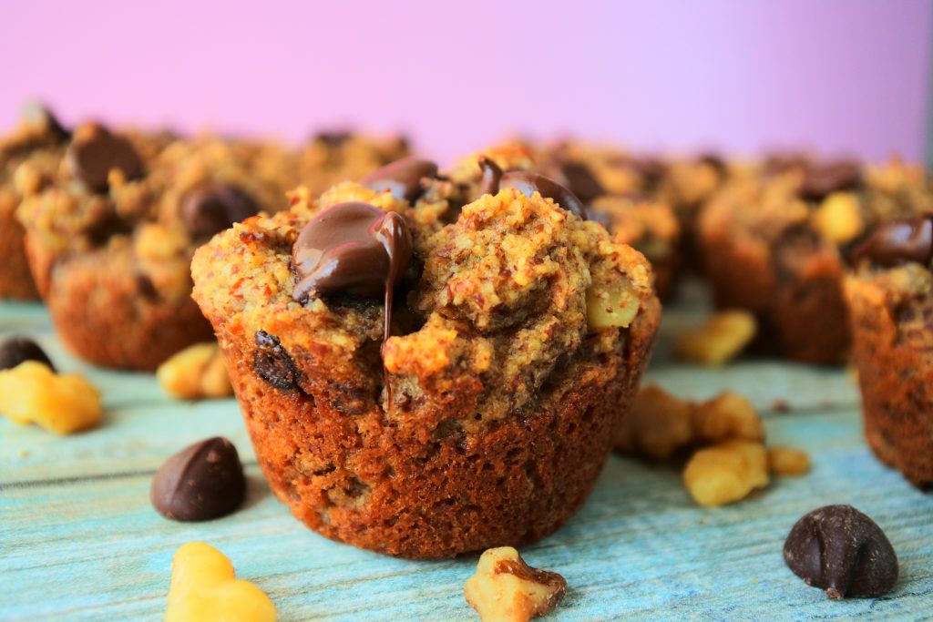 A close up image of a banana nut dark chocolate muffins surrounded by dark chocolate chips and walnuts