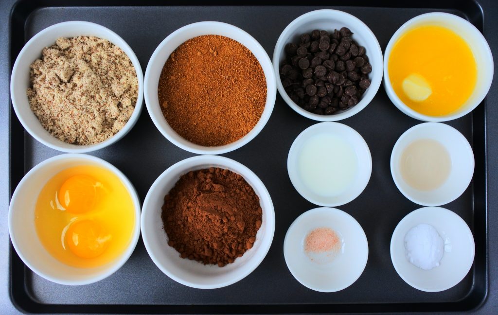 An overhead image of a tray of bowl containing the following ingredients: almond flour, eggs, coconut sugar, cocoa powder, dark chocolate chips, melted butter, a splash of milk, vanilla extract, salt and baking powder.