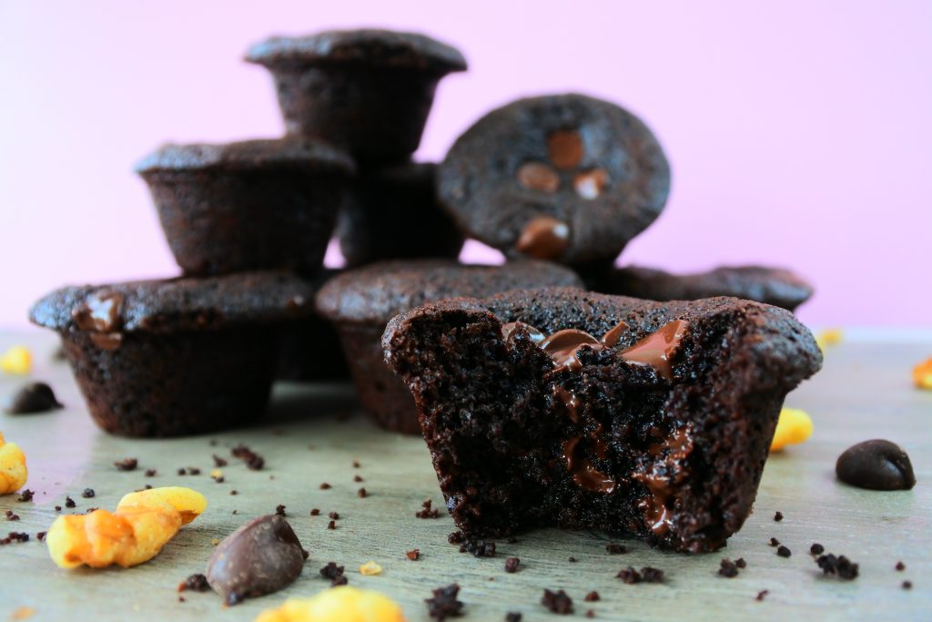 A head on, close up image of a mini muffin brownie surrounded by crumbs, chopped walnuts and dark chocolate chips with a pile of mini brownie muffins in the background.
