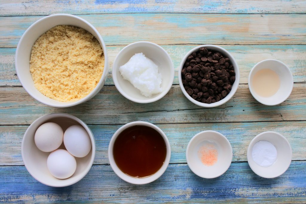 An overhead image of bowls of ingredients for chocolate chip muffins which include blanched almond flour, coconut oil, eggs, honey, salt, baking soda, vanilla extract and morsels of dark chocolate chips.