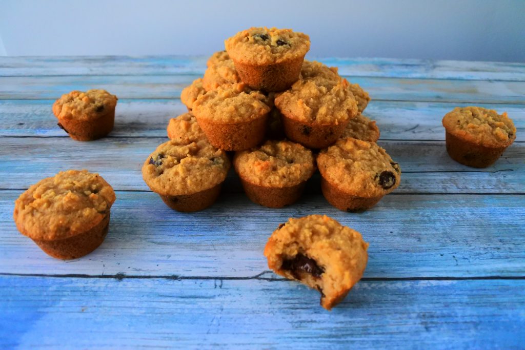 An angled image of a pyramid of chocolate chip mini muffins surrounded by four mini muffins one of which has had a bite taken out of it.