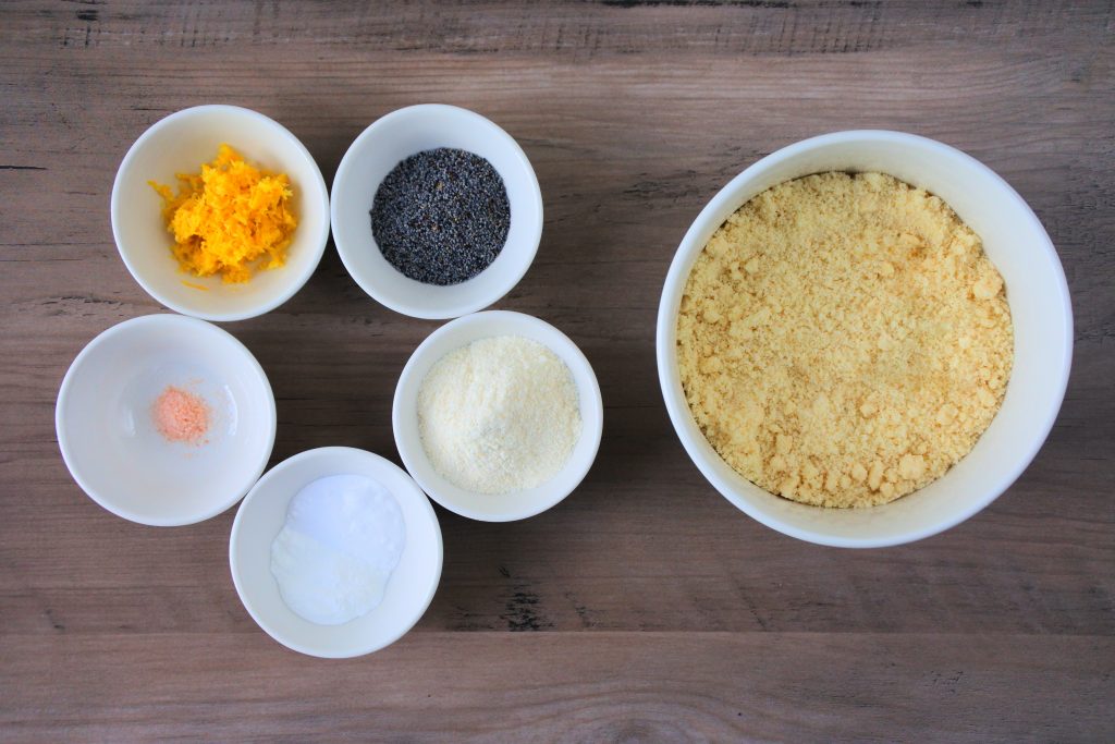 An overhead image of bowls of ingredients including blanched almond flour, coconut flour, baking powder and baking soda, salt, poppy seeds, and lemon zest