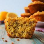 A head-on image of a close up of a split lemon poppyseed muffin with a plate of muffins and fresh lemons in the background