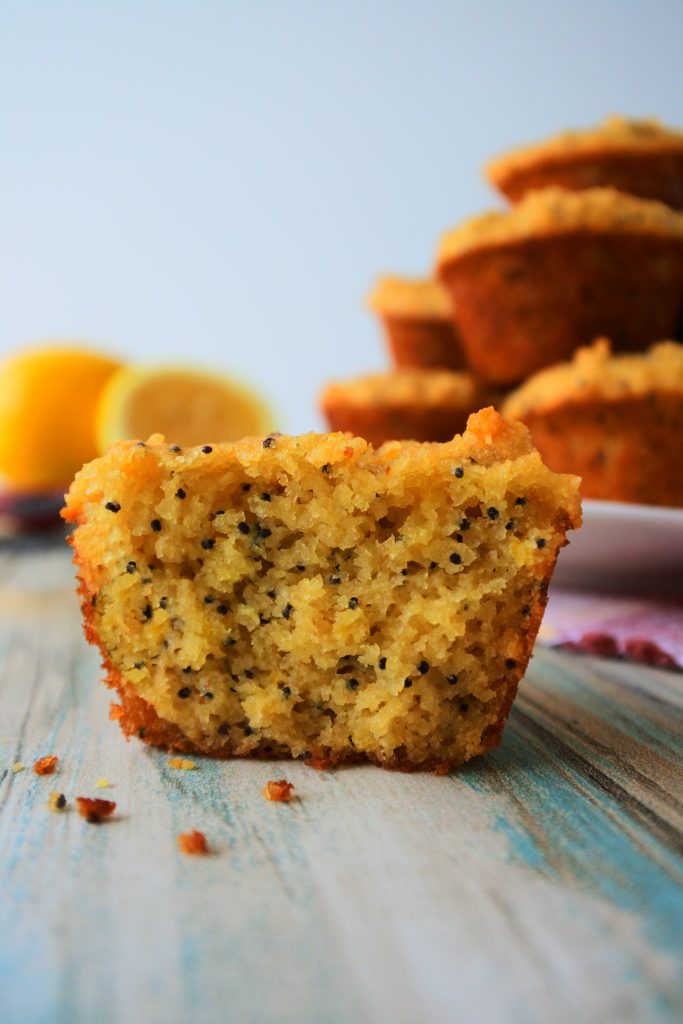 A head-on image of a close up of a split lemon poppyseed muffin with a plate of muffins and fresh lemons in the background