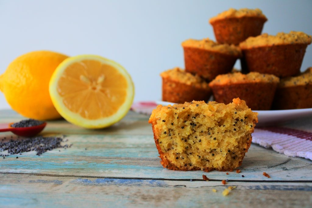 A head-on image of a close up of a split lemon poppyseed muffin with a plate of muffins, fresh lemons, and a teaspoon measure of poppyseeds in the background