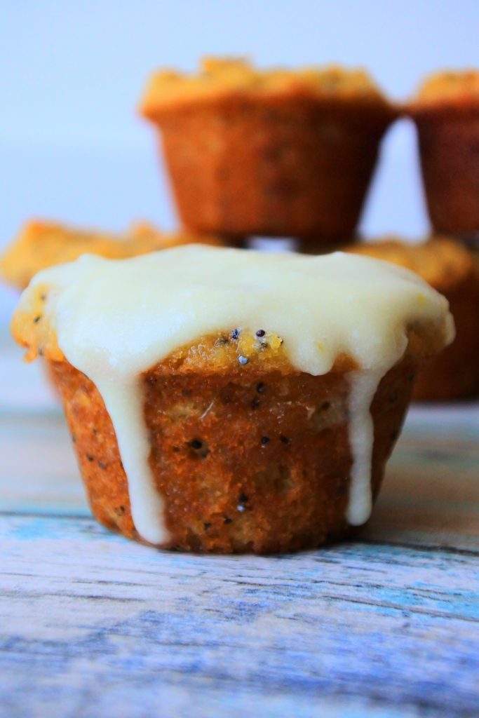 A vertical close up image of a mini lemon poppyseed muffin covered in a glaze with a stack of mini muffins in the background