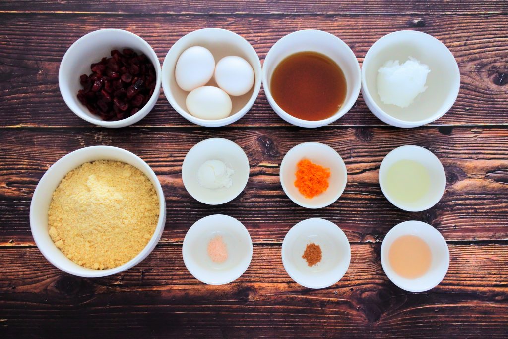 An overhead image of bowls of ingredients including dried cranberries, almond flour, eggs, maple syrup, coconut oil, baking powder, salt, nutmeg, vanilla extract, orange extract and orange zest.