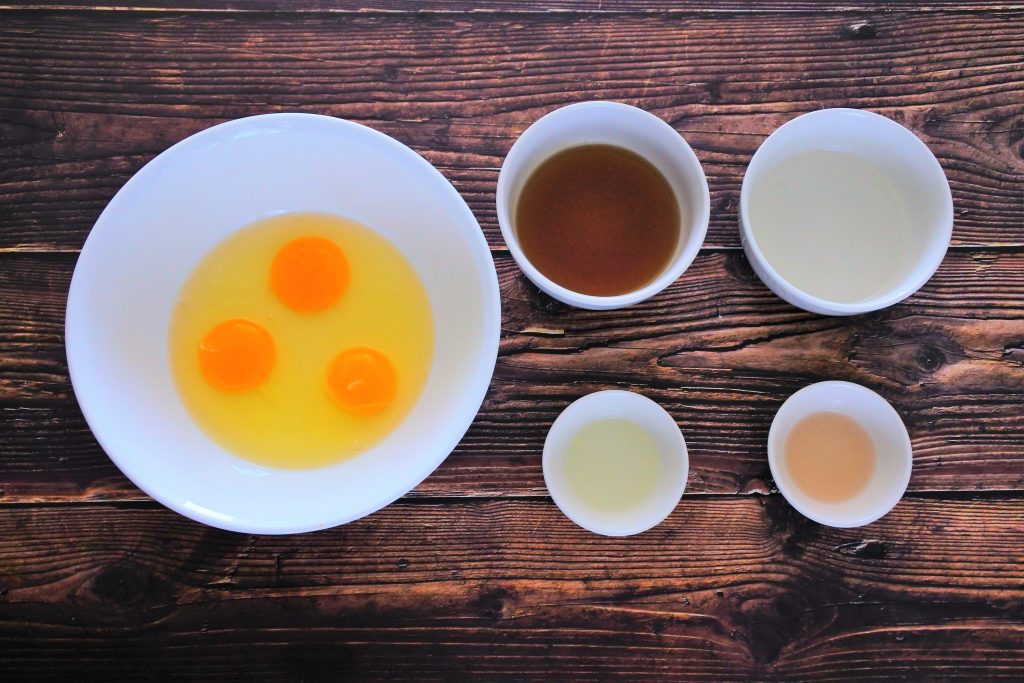 An over head image of bowls containing ingredients including cracked eggs, maple syrup, coconut oil (melted), orange extract and vanilla extract.
