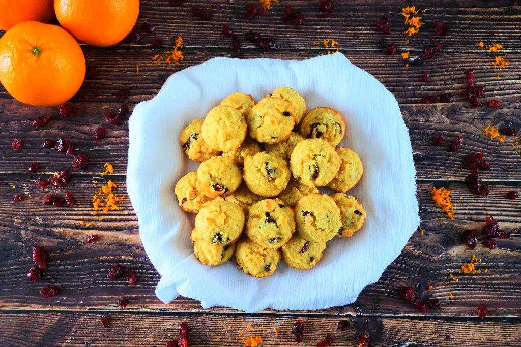 An overhead image of a pile of orange cranberry mini muffins on a white cloth surrounded by dried cranberries and orange zest with three whole oranges in the top left corner