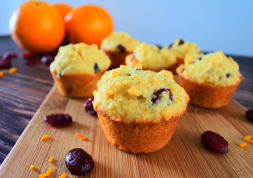 A close up image of some orange cranberry mini muffins on a wooden plank with dried cranberries and orange zest in the foreground and whole oranges in the background