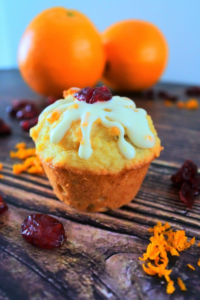 A close up image of a mini orange cranberry muffin topped with a cream cheese glaze and a dried cranberry surrounded by dried cranberries and orange zest with two oranges in the background