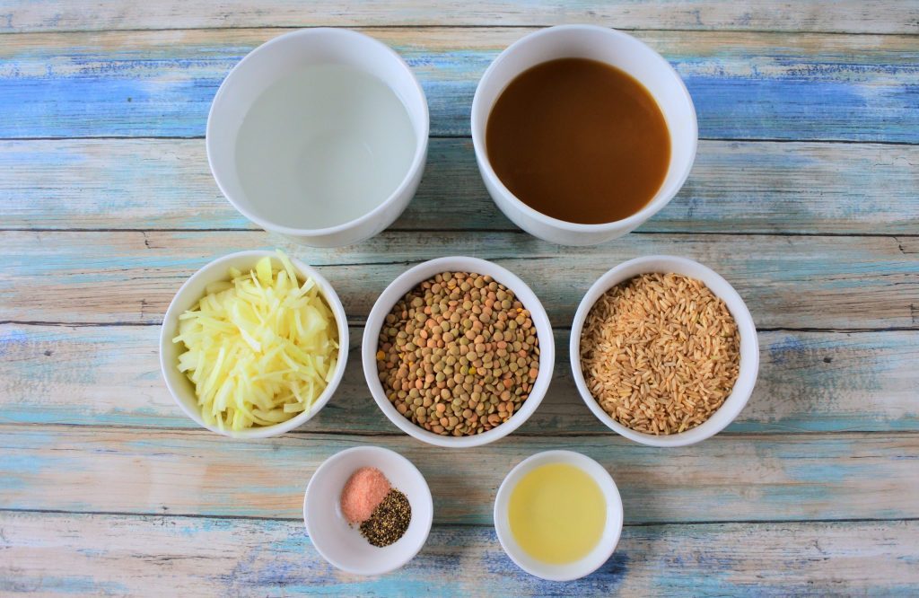 An overhead image of bowls of ingredients for mujadara including water, broth, sliced onions, dried lentils, brown rice, salt and pepper and extra light olive oil