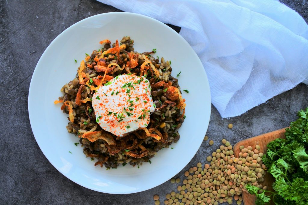 Am overhead image of a plate of mujadara topped with greek yogurt and crispy onions with white cloth next to a board with fresh herbs and dried lentils to the side