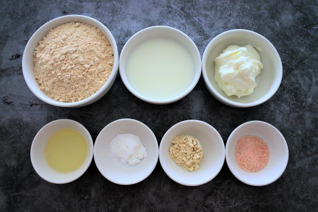 An overhead image of bowl of ingredients for naan including whole wheat flour, milk, yogurt, oil, baking powder, vital wheat gluten and salt.