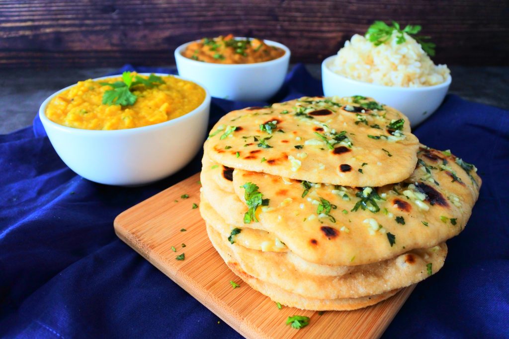 A close up image of a stack of garlic naan on a board with bowls of curry, dhal and rice in the background