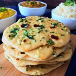A close up vertical image of a stack of garlic naan on a board with bowls of curry, dhal and rice in the background