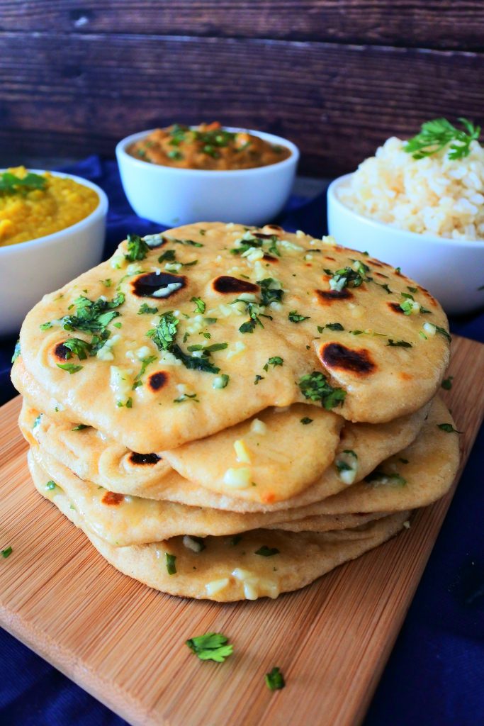 A close up vertical image of a stack of garlic naan on a board with bowls of curry, dhal and rice in the background