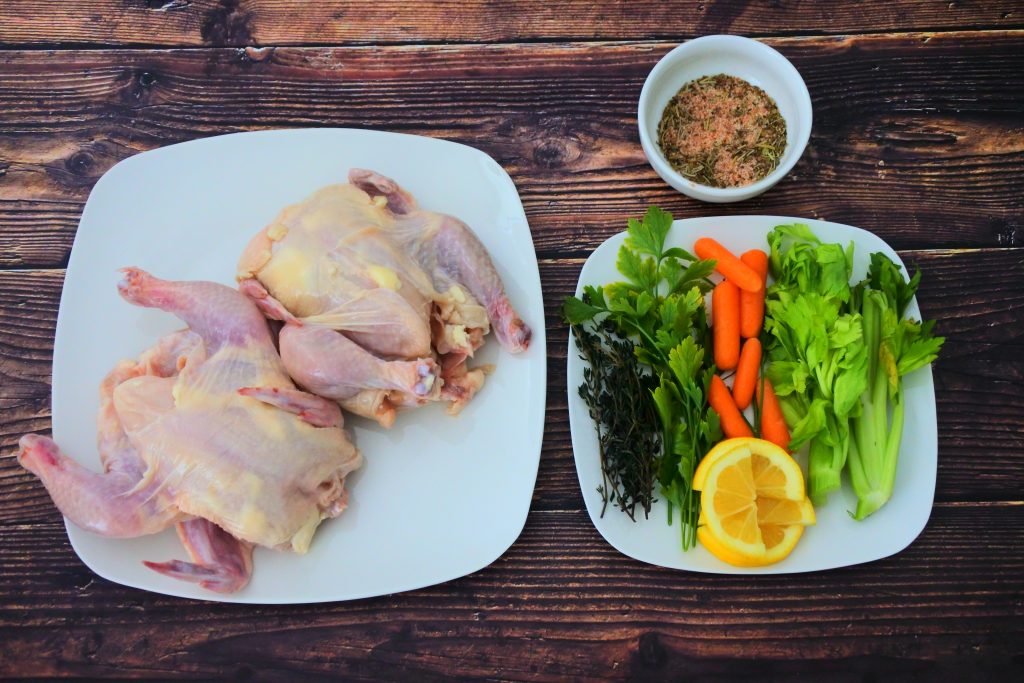 An overhead image of two cornish game hens with a bowl of spice rub next to a plate of herbs and vegetables for stuffing