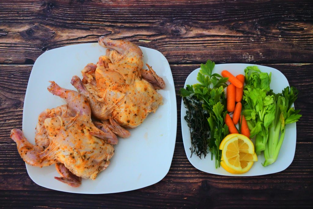 An overhead image of two cornish game hens with a rub next to a plate of herbs and vegetables for stuffing