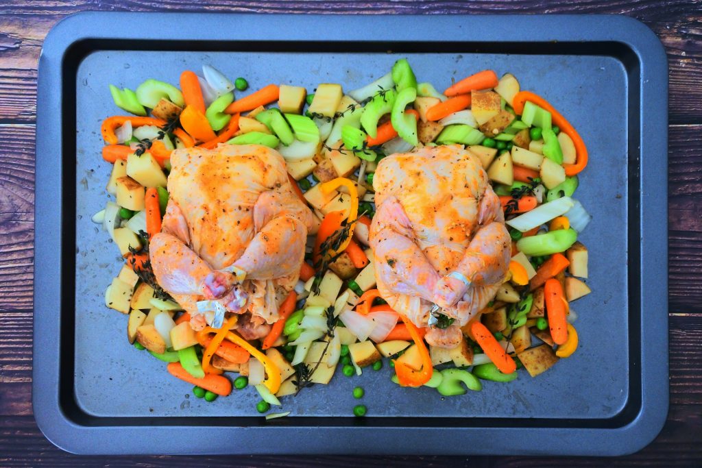 An overhead image of a tray of vegetables with two prepped cornish game hens on top