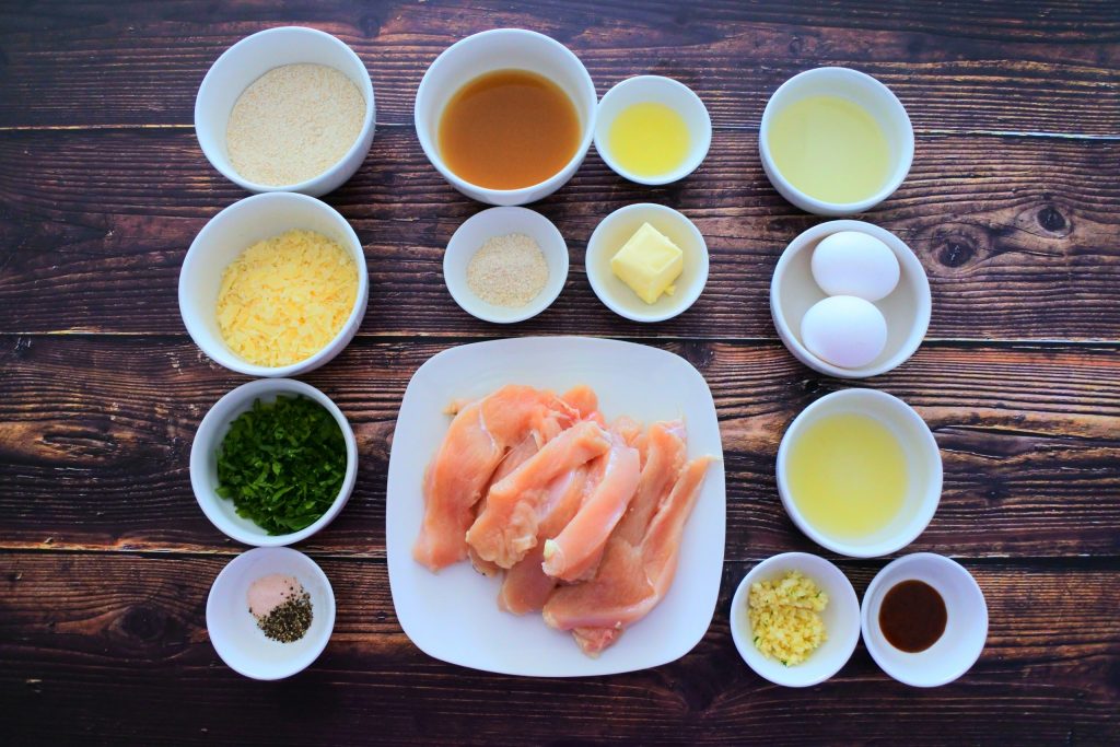 An overhead image of ingredients for herb crusted parmesan chicken including whole wheat flour, shredded paremensan cheese, herbs, salt, pepper, garlic, hot sauce, lemon juice, eggs, oil, and chicken breast cut into strips. And for a simple sauce, chicken bone broth, lemon juice, whole wheat flour and butter.