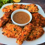 An angled image of a plate of herb crusted parmesan cutlets with a small dish of lemony gravy on a plate next to fresh herbs and a lemon half