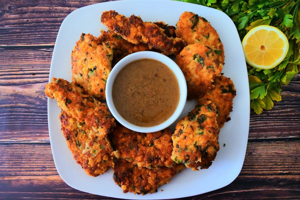 An overhead image of a plate of herb crusted parmesan cutlets with a small dish of lemony gravy on a plate next to fresh herbs and a lemon half