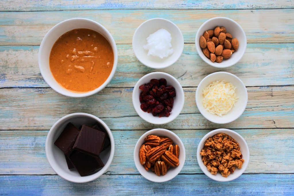 An overhead image of dishes of peanut butter, coconut oil, dark chocolate, almonds, dried cranberries, shredded coconut, pecans, and granola