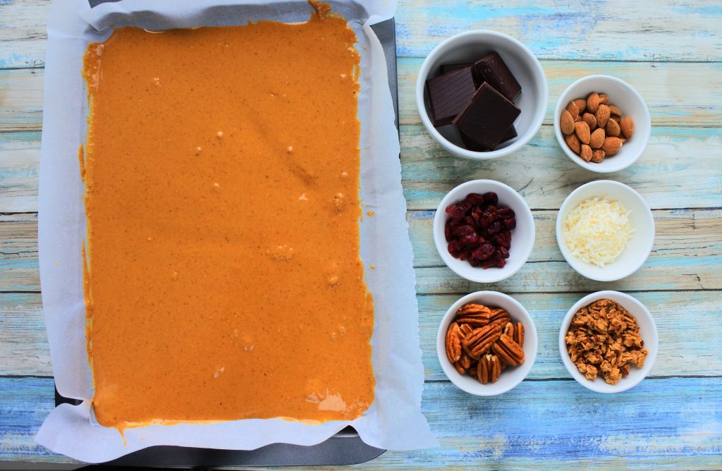 An overhead image of a tray of peanut butter with dishes of dark chocolate, almonds, dried cranberries, shredded coconut, pecans, and granola