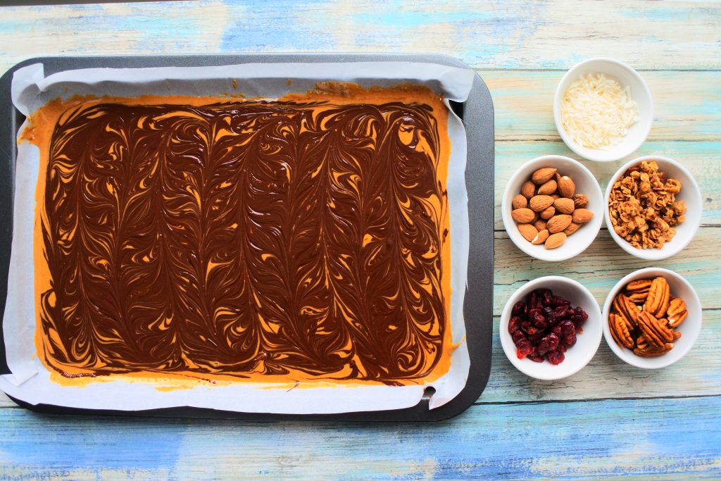 An overhead image of a tray with peanut butter with dark chocolate swirled in with small dishes filled with various toppings including shredded coconut, almonds, granola, dried cranberries, and pecans