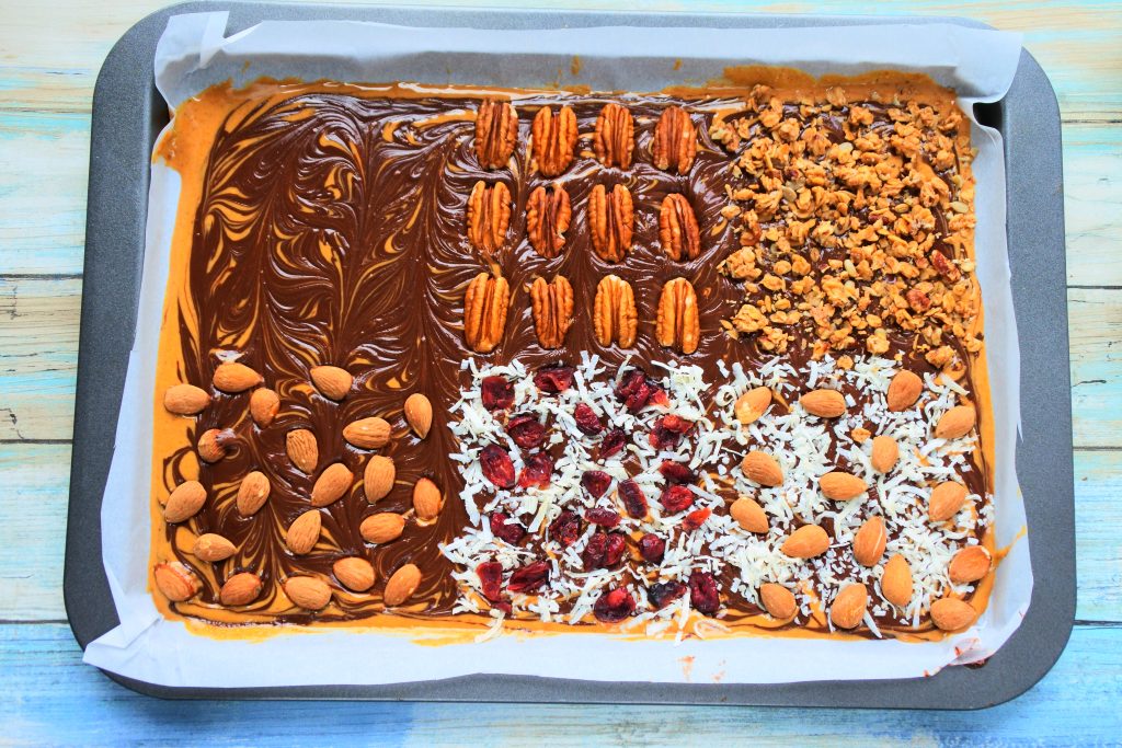 An overhead image of a tray of peanut butter and chocolate bark topped with various toppings included pecans, granola, almonds, shredded coconut and dried cranberries, and shredded coconut and almonds