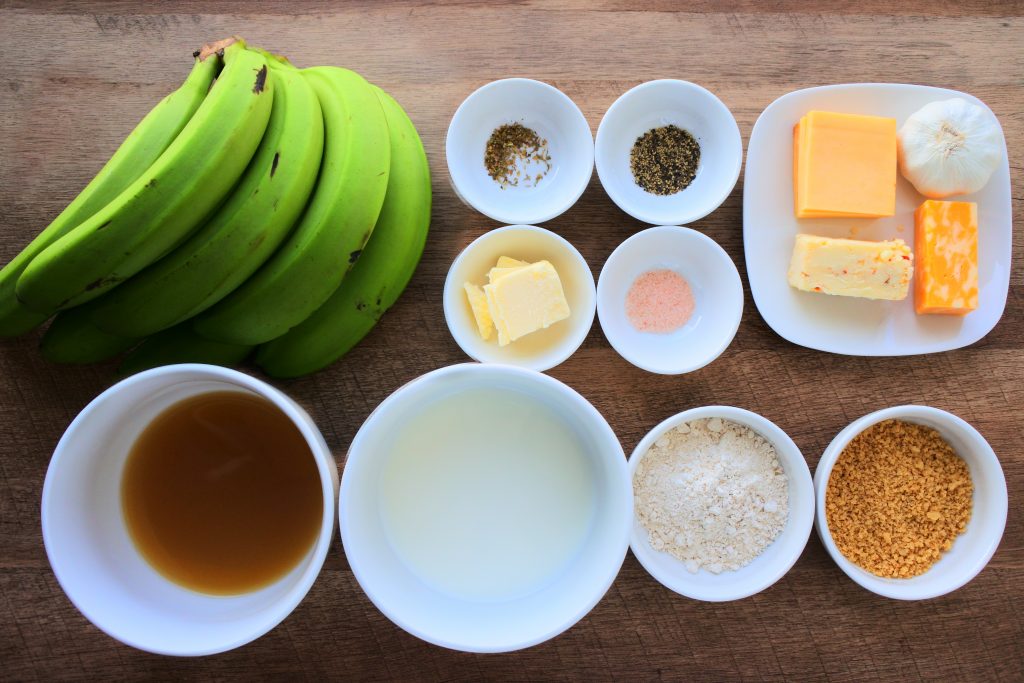 An overhead image of plates and dishes of ingredients for green banana pie including: green bananas, chicken broth, milk, whole wheat pastry flour, whole-wheat breadcrumbs, butter, salt, pepper, dried oregano, garlic, and cheeses (I used smoked Gouda, jalapeno cheddar and a marbled mozzarella/cheddar mix)
