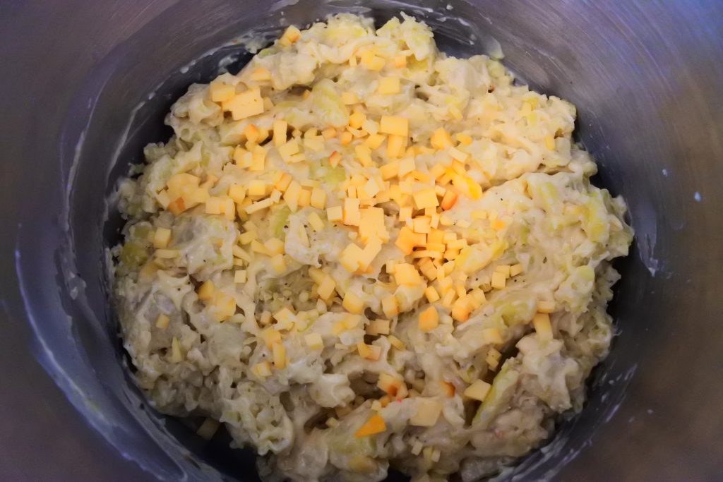 An overhead image of green bananas mixed with a cheesy roux sauce with small cubes of cheese on top