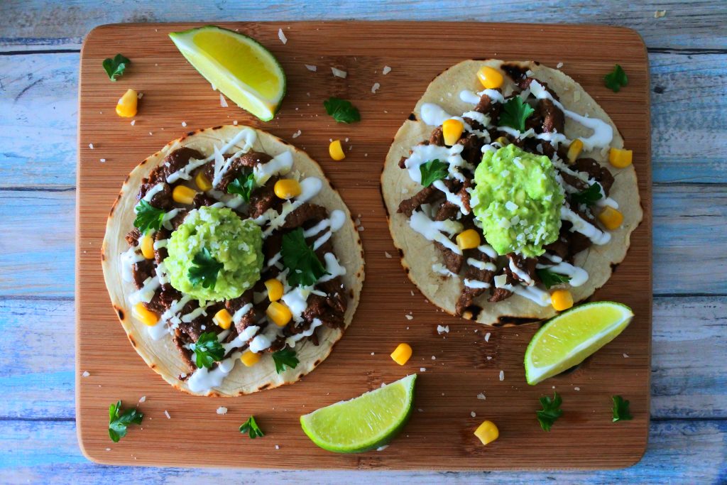 An overhead image of two corn tortillas topped with pineapple-soy marinated beef, crema, guacamole, corn, and cheese on a wood board