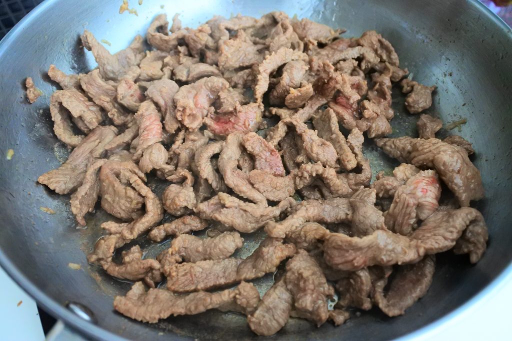 An angled image of thinly sliced marinaded beef cooking in a pan